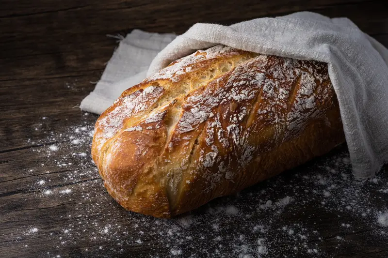 Freshly baked rustic loaf of bread covered with cloth, dusted with flour on wooden table