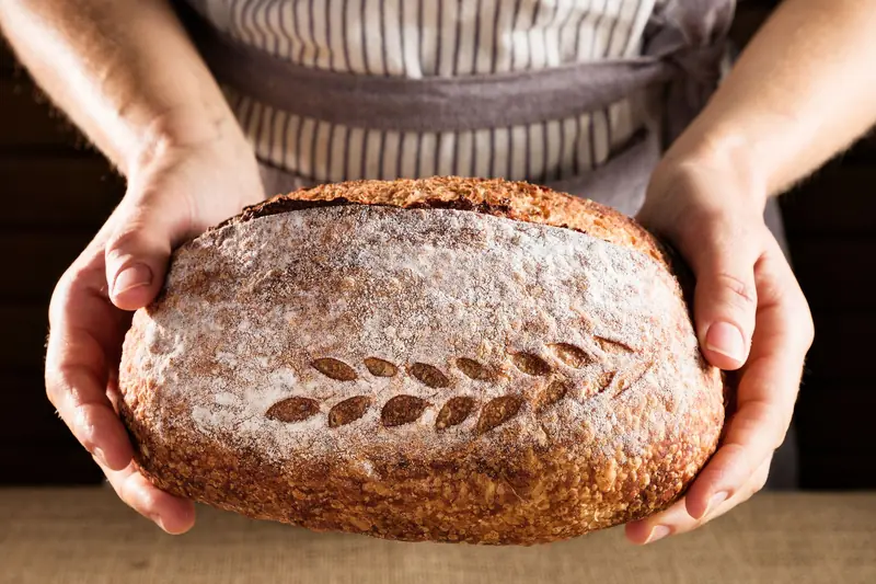 Rustic sourdough loaf with a decorative pattern, held by hands in a warm, artisanal kitchen setting