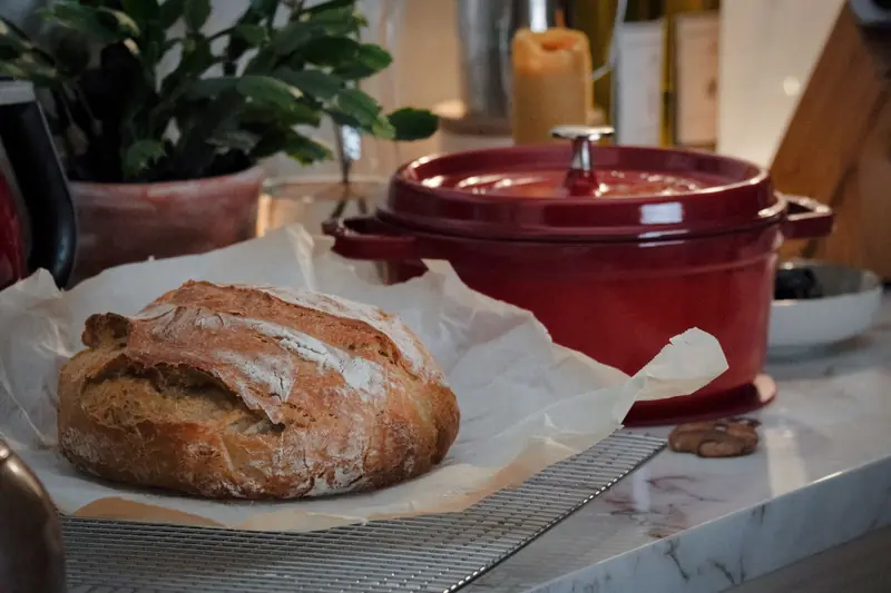 Close-up of a rustic loaf of bread on a kitchen counter with a red Dutch oven nearby